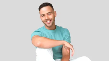 A man in a teal shirt and white pants smiles against a plain background.