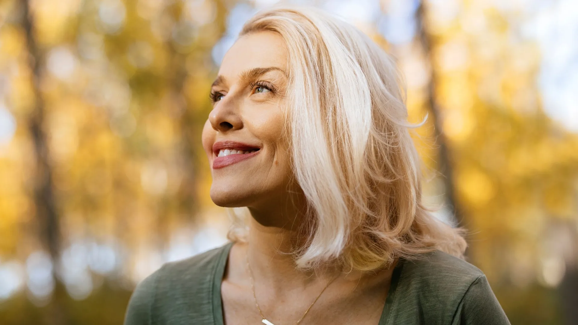 A woman with blonde hair smiling outdoors, with a blurred autumn background.