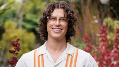 A person smiling outdoors, wearing glasses and a shirt with orange stripes, surrounded by lush greenery and red plants.