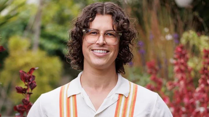 A person smiling outdoors, wearing glasses and a shirt with orange stripes, surrounded by lush greenery and red plants.