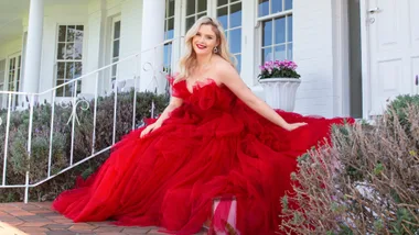 A woman in a flowing red gown sitting on steps in front of a white building with flowers around.