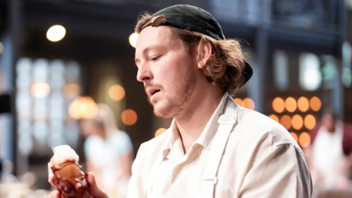 Man in a white apron and black cap, focused on cooking in a dimly lit kitchen, holding a food item.