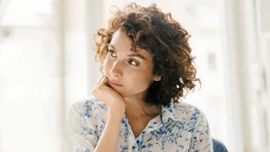 Young woman with curly hair, resting her chin on her hand, wearing a floral blouse, gazes thoughtfully out a window.
