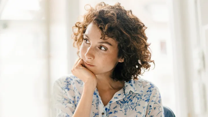 Young woman with curly hair, resting her chin on her hand, wearing a floral blouse, gazes thoughtfully out a window.