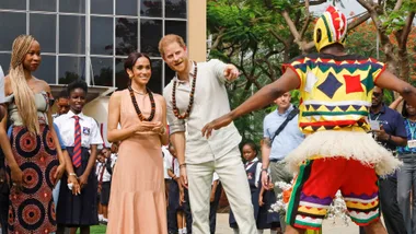 Group of people, including a man and woman in necklaces, watch a person in colorful attire, outdoors with children.