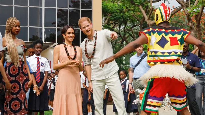 Group of people, including a man and woman in necklaces, watch a person in colorful attire, outdoors with children.