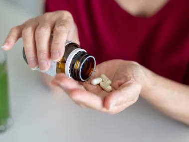 Person pouring white capsules from a brown bottle into their hand, with a glass of green liquid nearby.