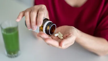 Person pouring white capsules from a brown bottle into their hand, with a glass of green liquid nearby.