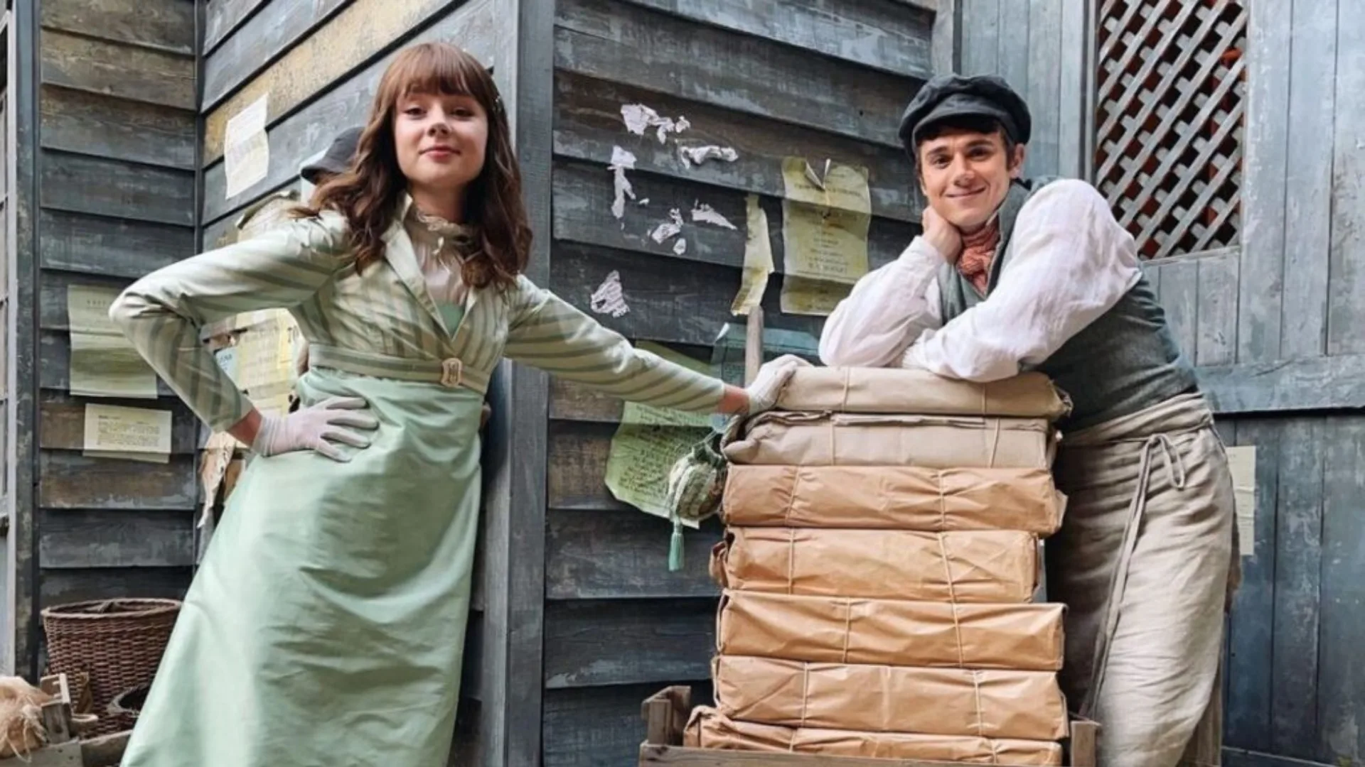 Two people in vintage attire, one leaning on a stack of wrapped packages, standing against a wooden wall with posters.