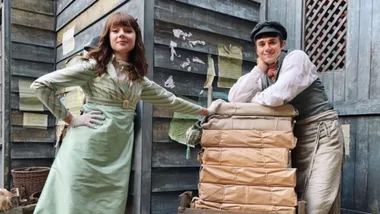 Two people in vintage attire, one leaning on a stack of wrapped packages, standing against a wooden wall with posters.