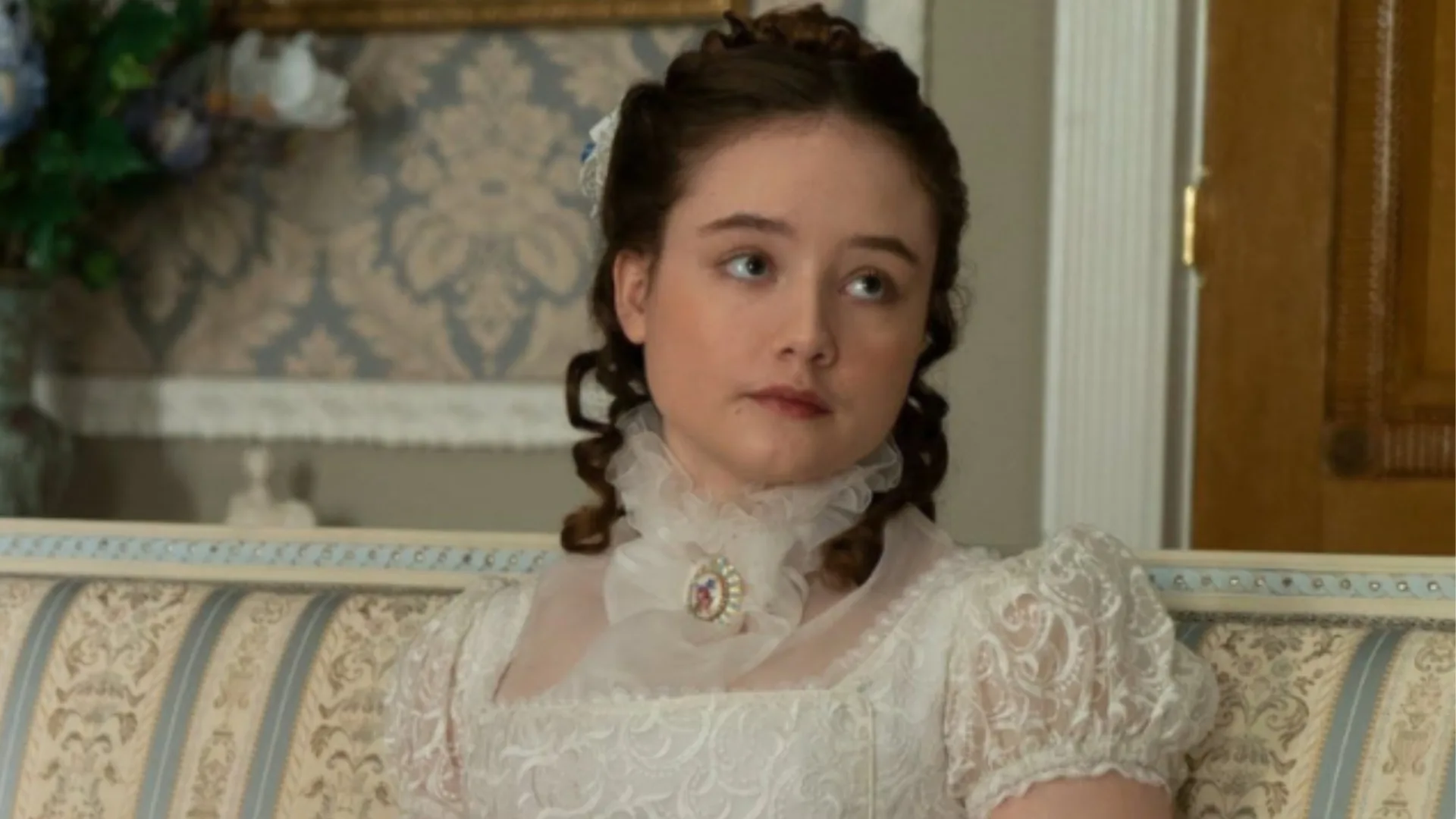Young woman in a white Victorian dress with lace, sitting on a patterned sofa in an ornate room.