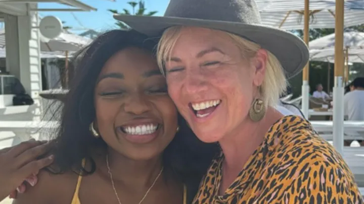 Two people smiling outdoors; one in a yellow outfit, the other in a leopard print shirt and hat.