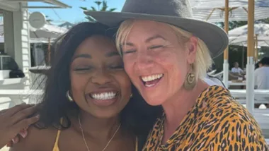 Two people smiling outdoors; one in a yellow outfit, the other in a leopard print shirt and hat.