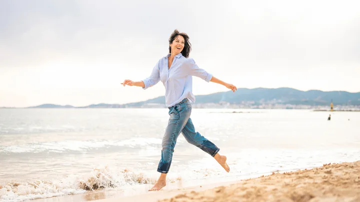 A woman in a light blue shirt and jeans joyfully runs along a sandy beach with waves in the background.