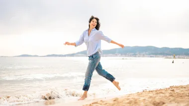 A woman in a light blue shirt and jeans joyfully runs along a sandy beach with waves in the background.