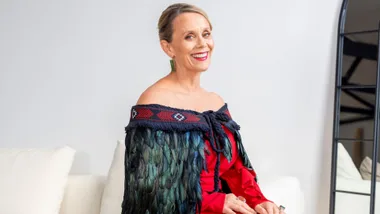 A woman in a feathered korowai and red dress smiles while sitting on a white couch.