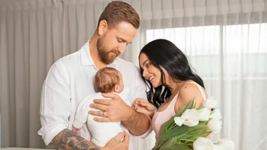 A couple lovingly holds their baby, with the mother holding a bouquet of white tulips, set within a cozy home environment.