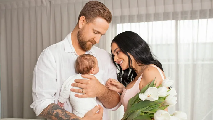 A couple lovingly holds their baby, with the mother holding a bouquet of white tulips, set within a cozy home environment.