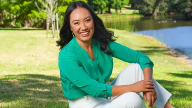 Woman in a green shirt and white pants sitting on grass by a pond, smiling with trees in the background.