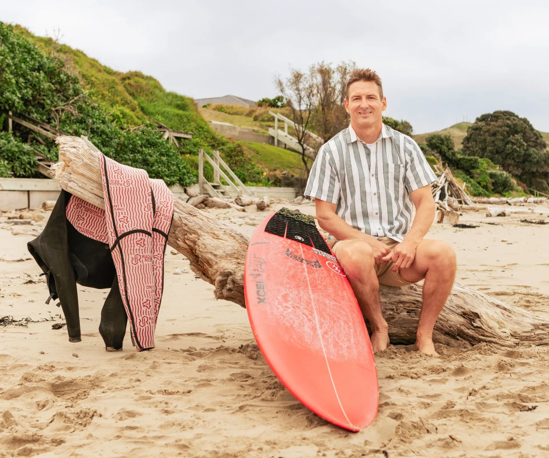 Maz Quinn sitting on a piece of driftwood on a beach beside his surf board and wetsuit