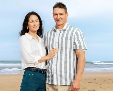 A woman and man stand together smiling on a beach with waves in the background.