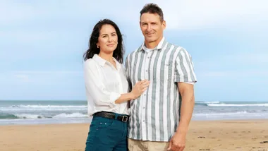 A woman and man stand together smiling on a beach with waves in the background.