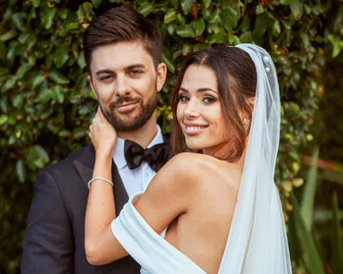 Couple posing on their wedding day; bride in white dress and veil, groom in black tuxedo, smiling in front of greenery.