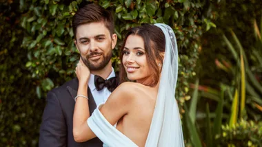 Couple posing on their wedding day; bride in white dress and veil, groom in black tuxedo, smiling in front of greenery.