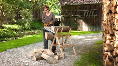 Person using an electric chainsaw to cut logs on a sawhorse outdoors, with stacked firewood nearby.