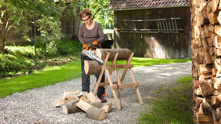 Person using an electric chainsaw to cut logs on a sawhorse outdoors, with stacked firewood nearby.