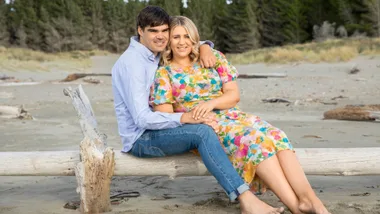 Couple sits on a beach log with trees in background; man in blue shirt, woman in floral dress, both smiling.