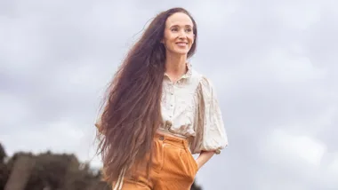 A woman with long hair smiles outdoors, wearing a beige blouse and orange trousers against a cloudy sky.