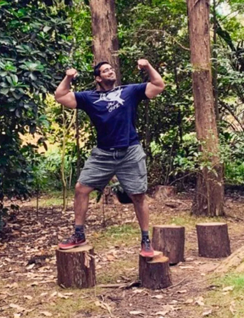 Suraj standing with each foot on a tree stump, flexing his biceps