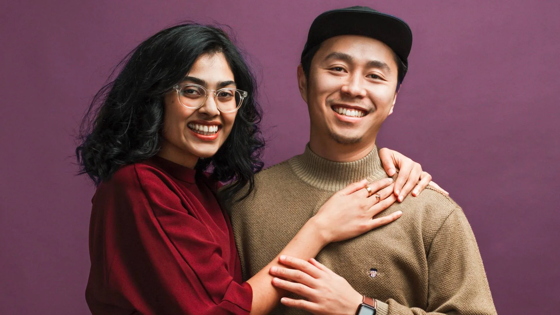 Ankita Sigh has her hands around Calvin Sang's neck, both smiling for the camera in front of a purple background