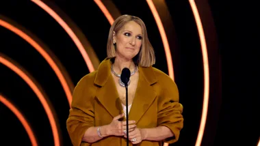 Celine Dion speaking on stage in a mustard coat with curved light patterns behind her.