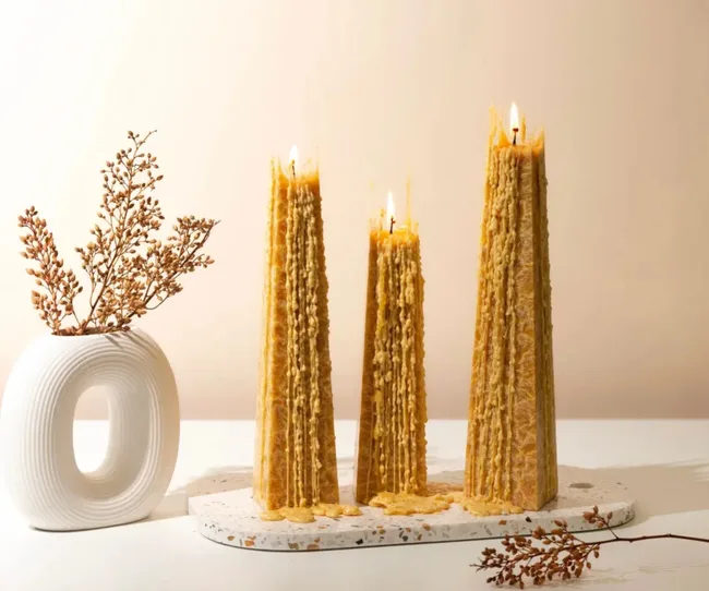 Three tall, textured candles burning beside a modern white vase with dried branches on a neutral background.