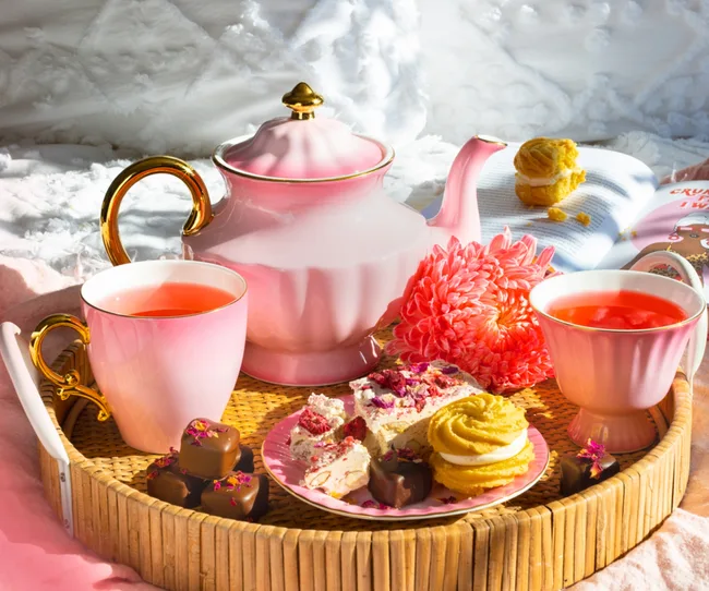 Pink tea set on a wicker tray with tea, desserts, and flowers in afternoon sunlight.