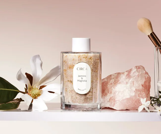 Circa Jasmine & Magnolia bath salts with a flower, pink crystal, and brush on a shelf.