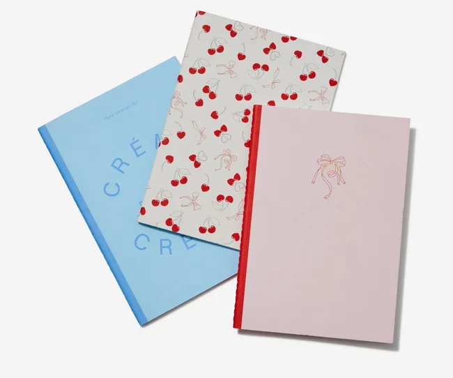 Three notebooks stacked: one blue with the word "CR&Eacute;A", one with cherry pattern, and one pink with a bow illustration.