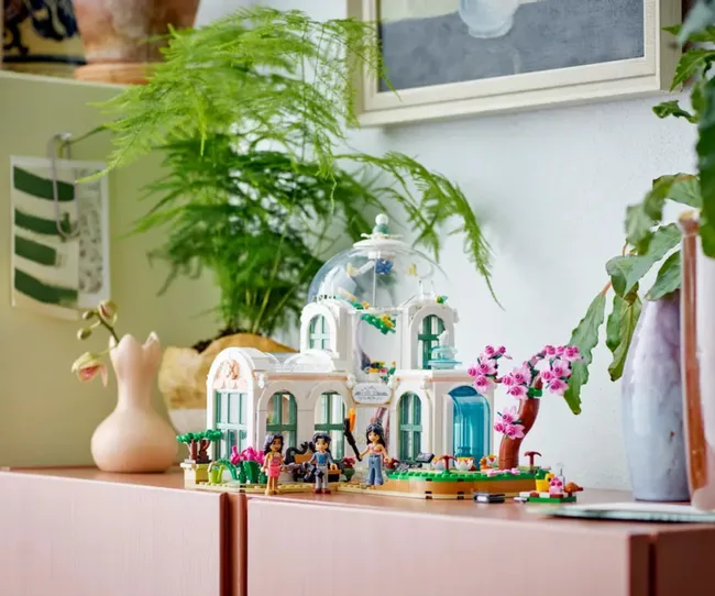 Lego botanical garden set with mini-figures displayed on a wooden cabinet, surrounded by plants and decor.