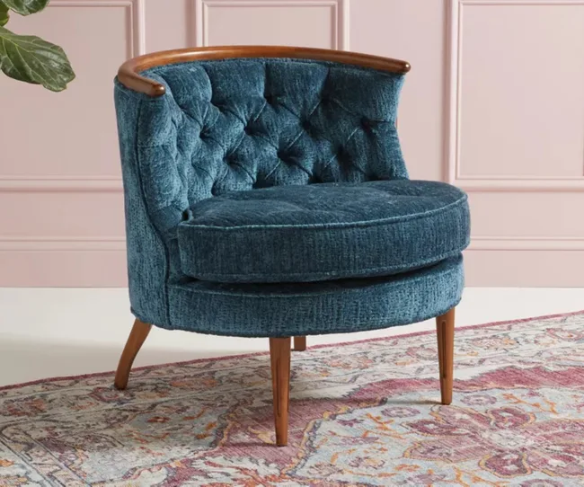 Blue tufted accent chair with wooden trim and legs on a patterned rug in a room with pink paneled walls.