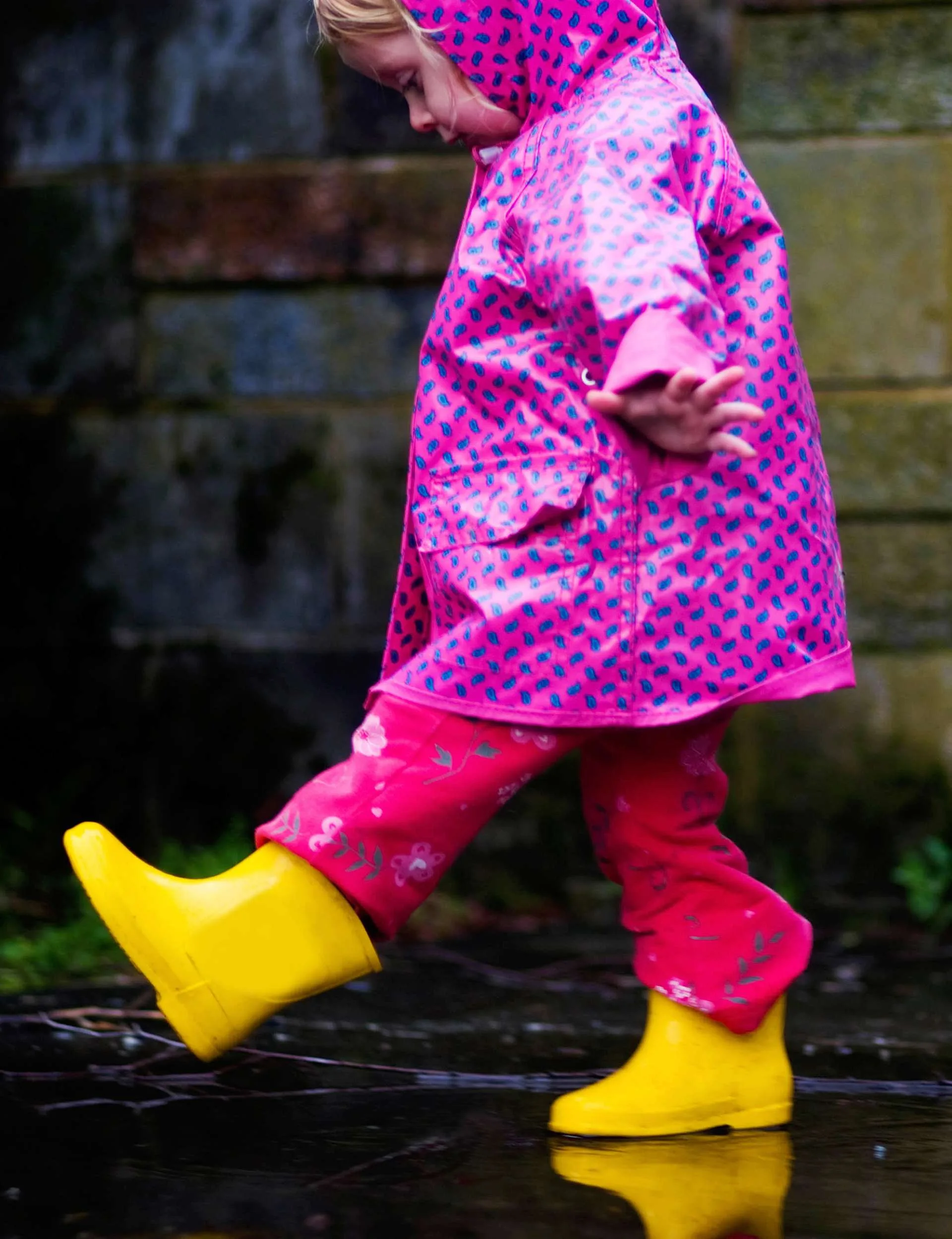 A toddler in a pink raincoat and yellow gumboots stomping in puddles