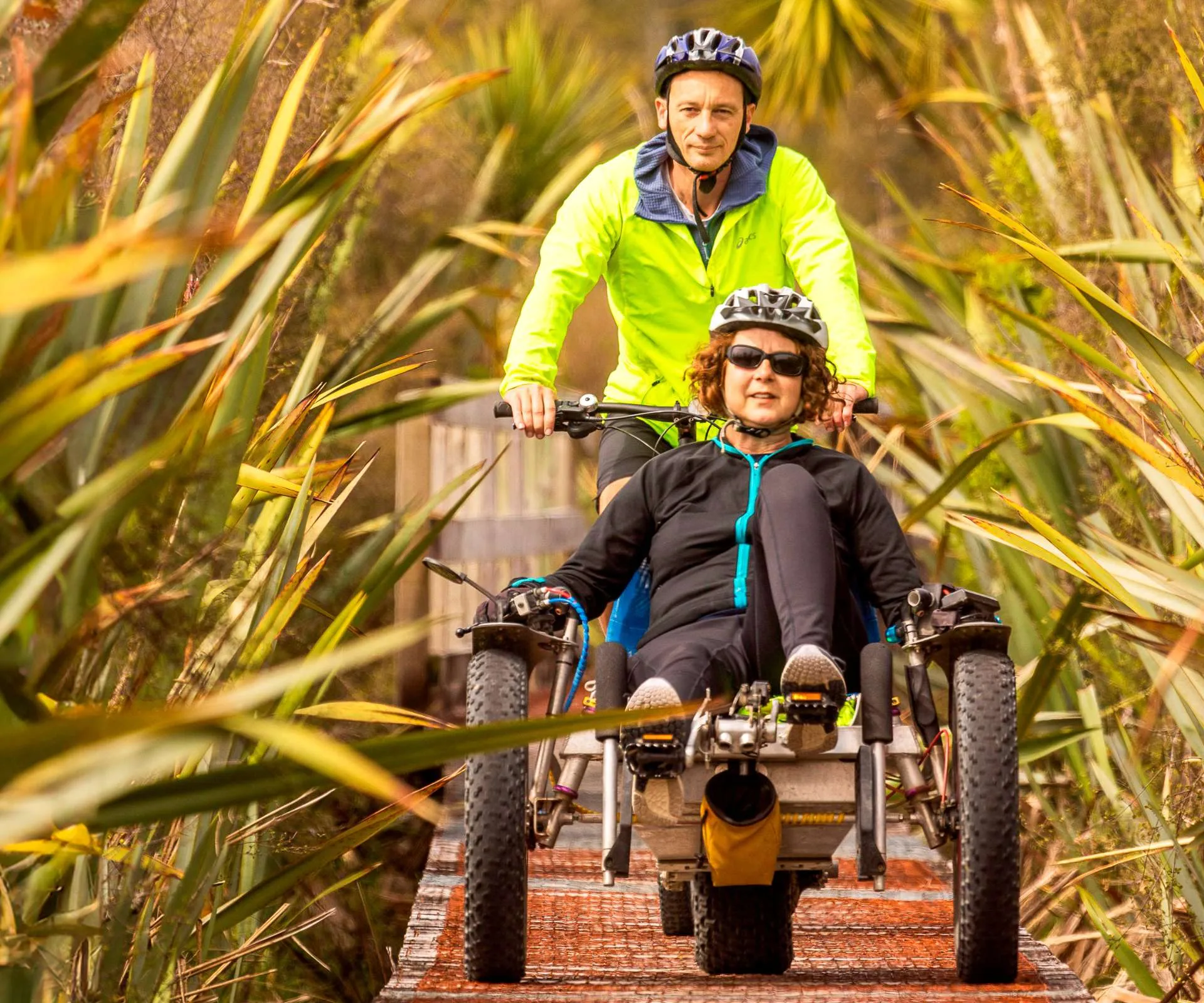 Dr Natalie in her wheelchair with husband Matt biking behind her on a wooden path surrounded by flax