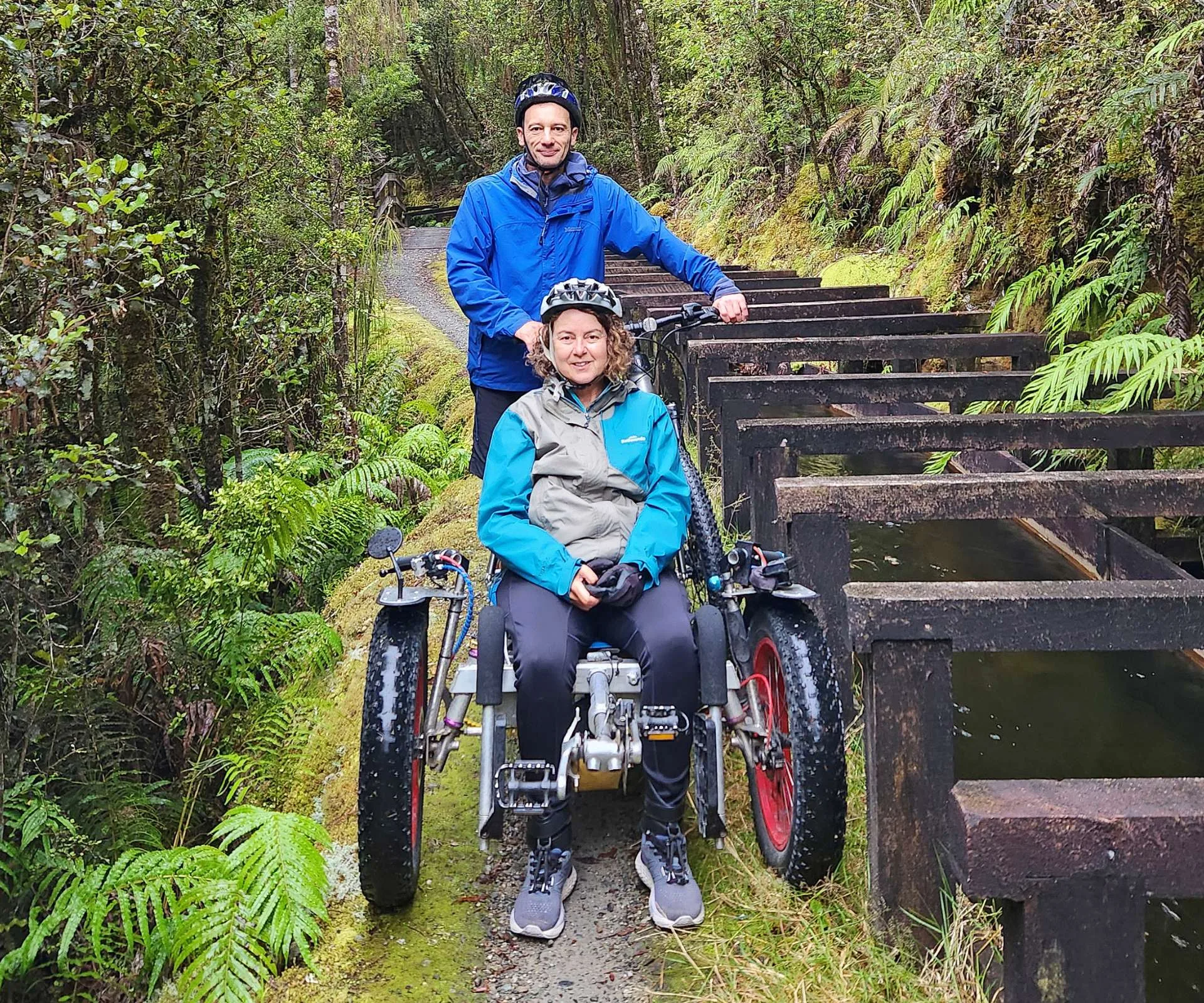Dr Natalie in her wheelchair with husband Matt standing with his bike behind her on a native track