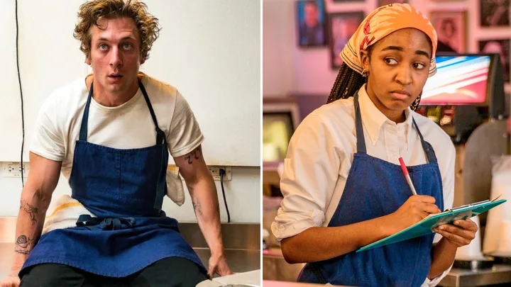 Two chefs in blue aprons, one seated looking up, the other holding a clipboard, appear focused and thoughtful.