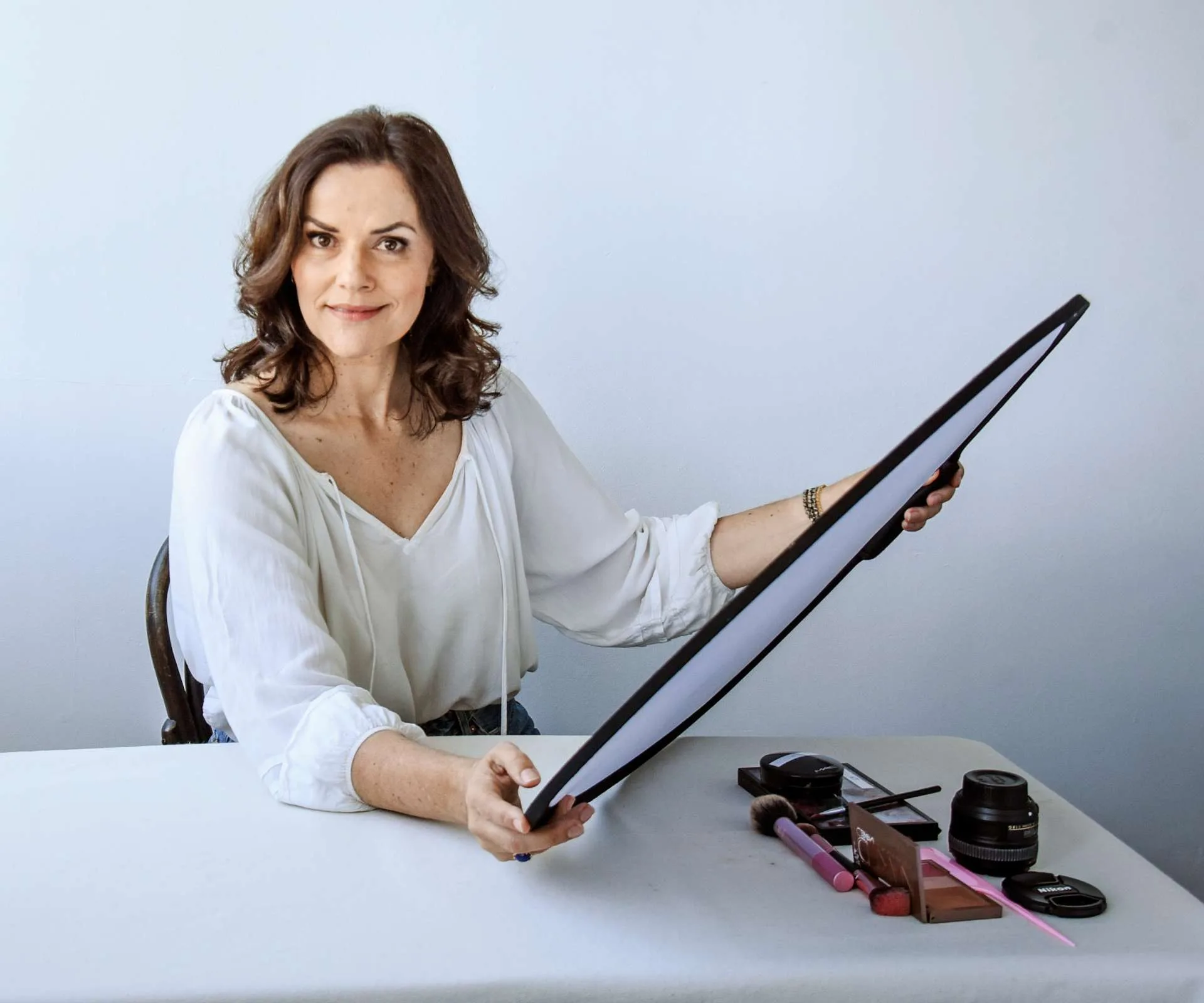 Amanda Billing holding a light sheet with makeup and a camera lens on the table in front of her