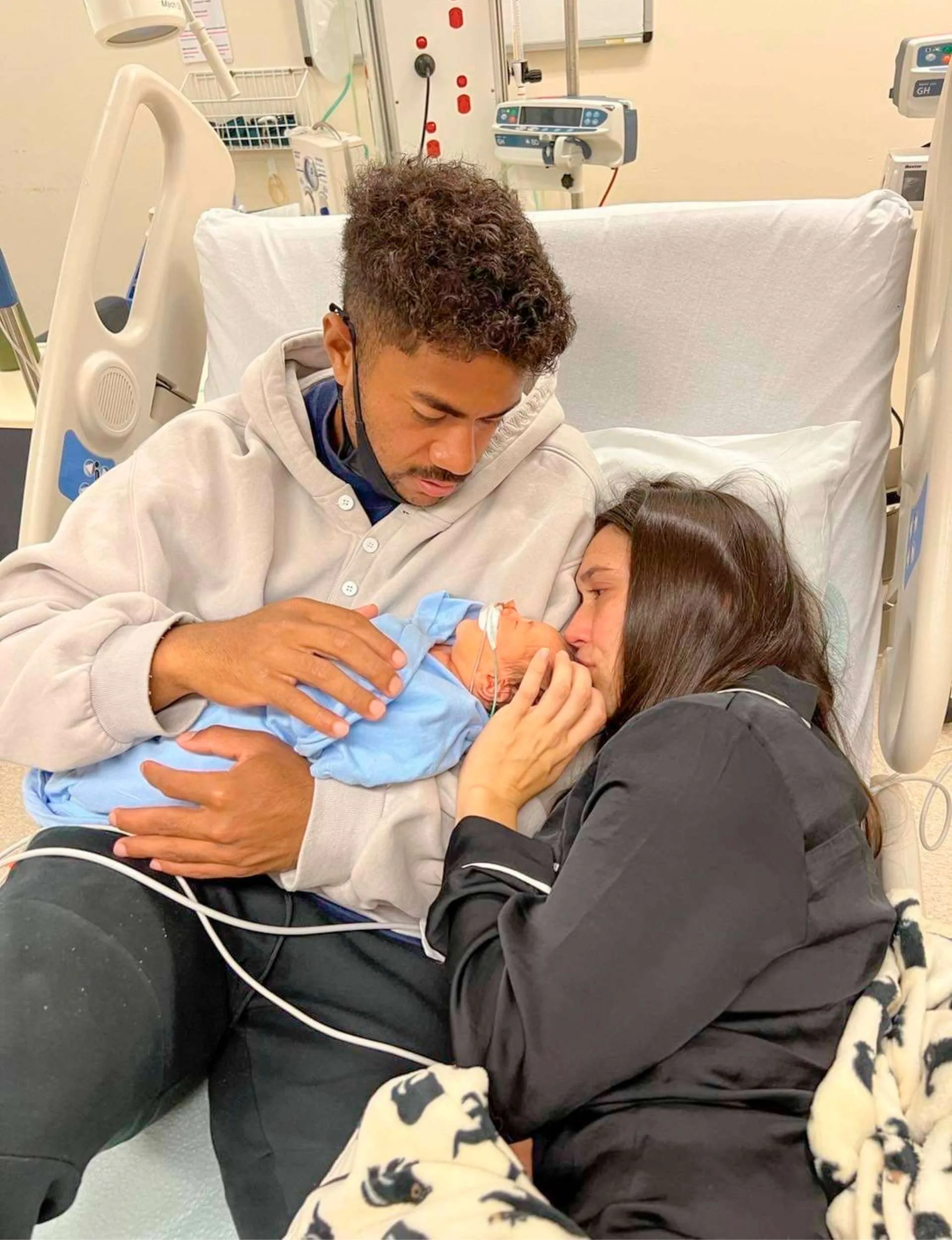 Emoni and Danielle laying in a hospital bed holding newborn Milla, while she's connected to tubes and wires