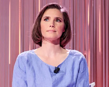 Amanda Knox sitting in front of a pink background with a mic attached to her top