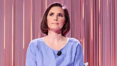 Amanda Knox sitting in front of a pink background with a mic attached to her top