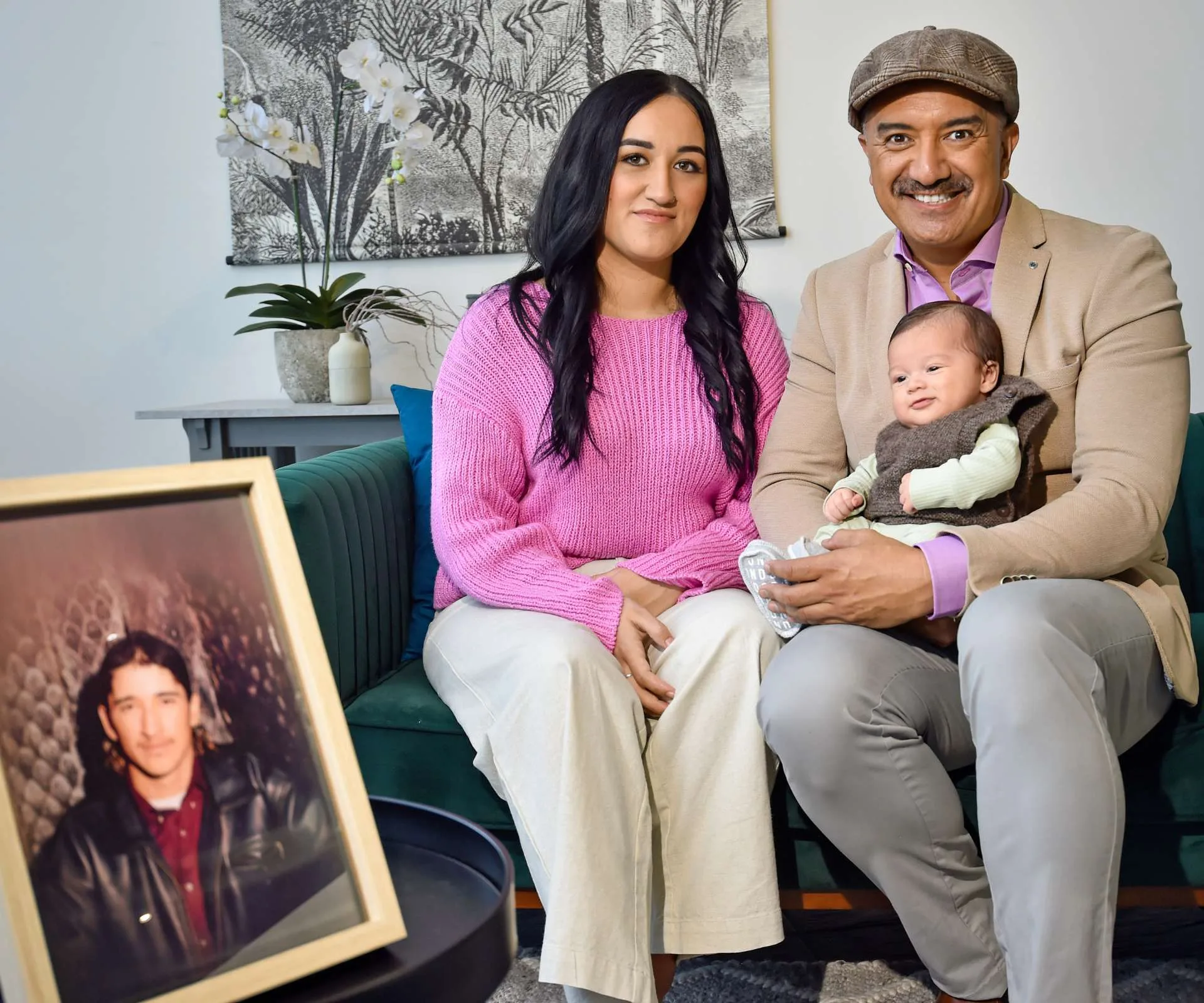 Mātai Smith holding baby Raekahu, sitting on a couch beside niece Samara and a framed photo of late brother Hakim.